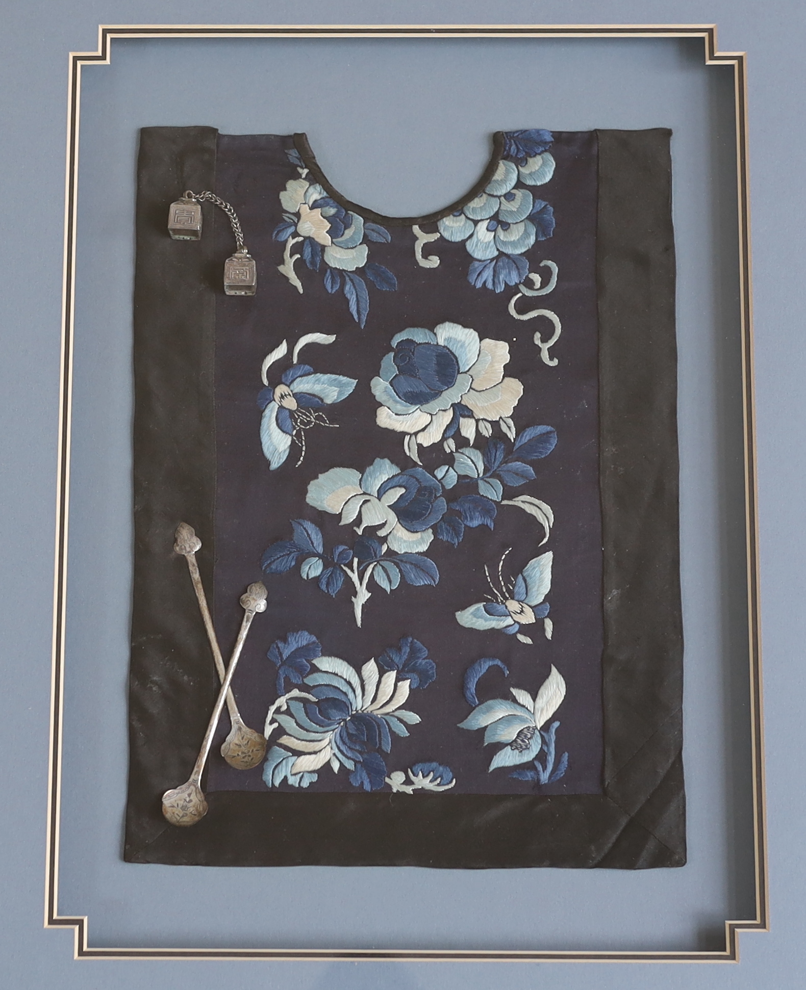 A late 19th century Chinese silk and blue floral embroidered baby’s bib, mounted with two white metal spoons and a rattle, together with a similar bib and spoons plus a decorative white metal mask mount, blue embroidery